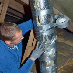 Man i blå tröja tejpar en isolerad ventilationsslang på en vind.