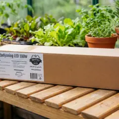 En kartong med växtbelysning LED 100W ligger på ett träbord bland plantor. Perfekt för att ge dina grönsaker extra ljus.