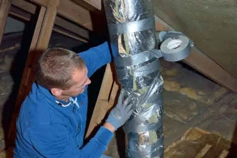 Man i blå tröja tejpar en isolerad ventilationsslang på en vind.