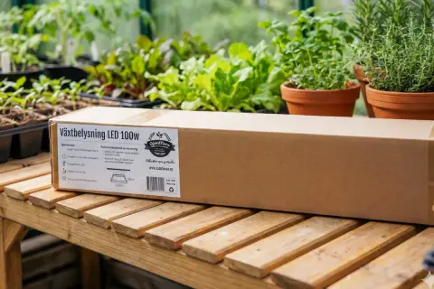 En kartong med växtbelysning LED 100W ligger på ett träbord bland plantor. Perfekt för att ge dina grönsaker extra ljus.