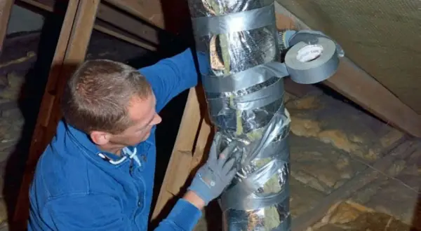 Man i blå tröja tejpar en isolerad ventilationsslang på en vind.