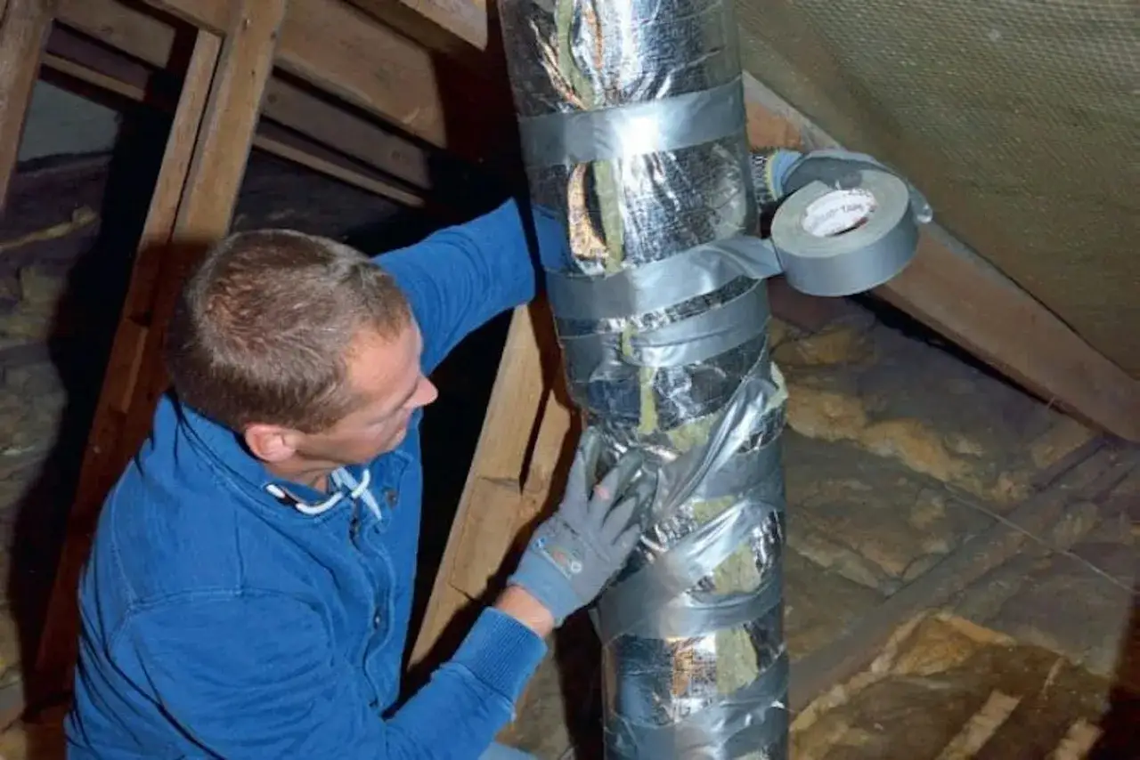 Man i blå tröja tejpar en isolerad ventilationsslang på en vind.