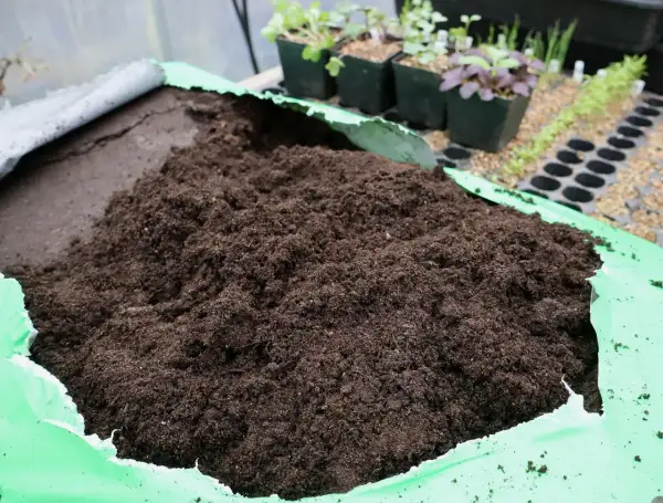 En stor hög med mörk blomjord ligger i en öppen grön plastpåse. I bakgrunden syns små plantor i krukor.